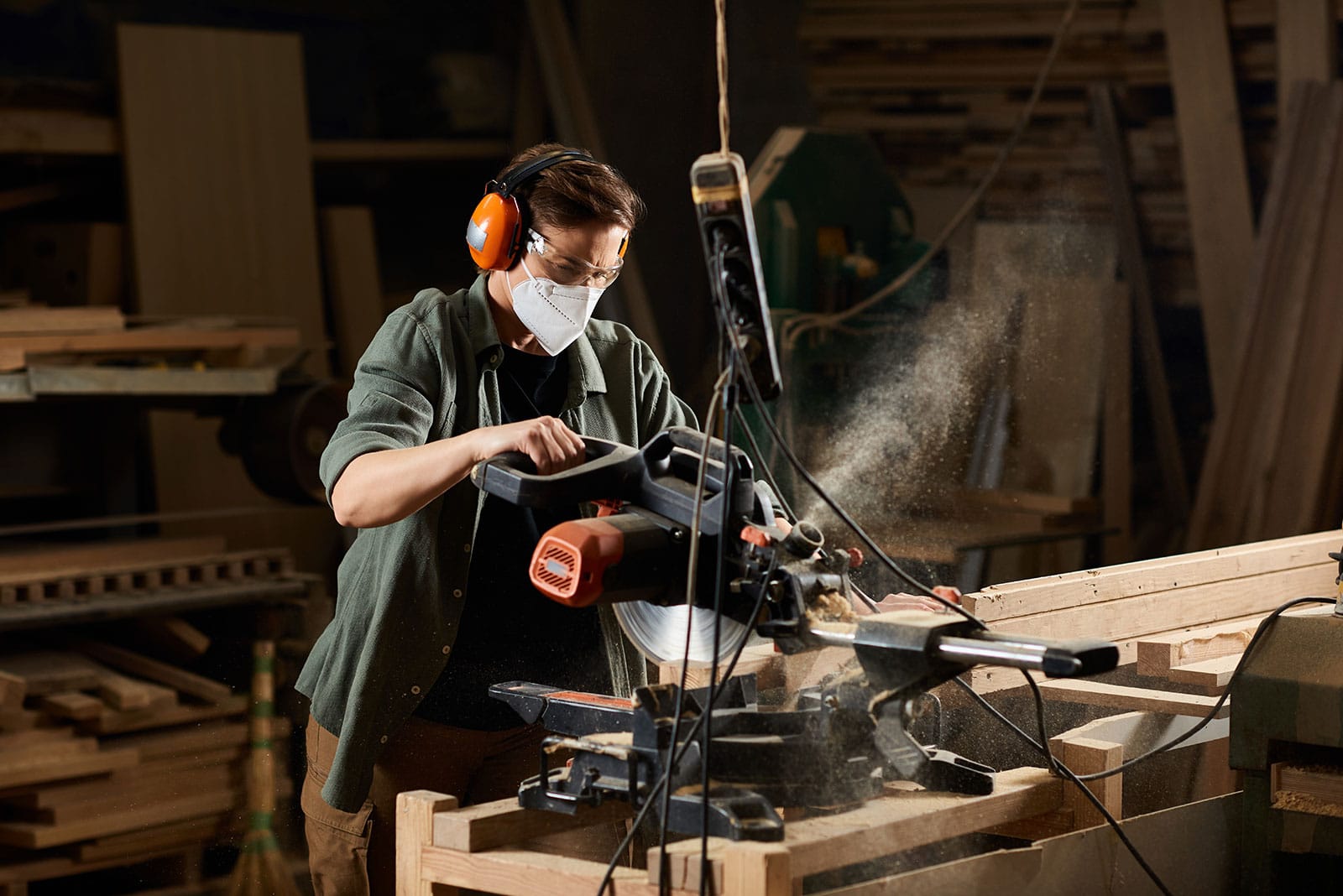 Worker operating a power saw in a woodworking shop, wearing safety equipment for protection.