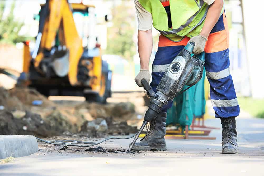 Construction worker with power drill on road site, wearing safety gear.