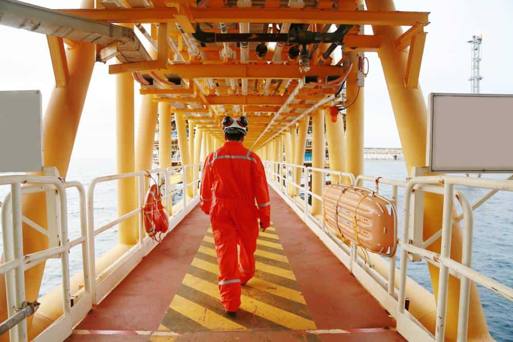 Offshore worker in red safety gear walking on an oil platform deck over water.