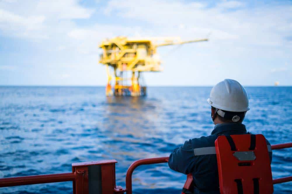 Offshore worker overlooking oil rig with health and safety gear in maritime environment.