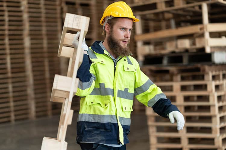 Utility worker in safety gear carrying wooden pallet in warehouse.