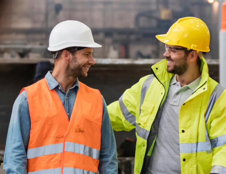 Two construction workers smiling and talking on site, wearing safety helmets and high-visibility jac.